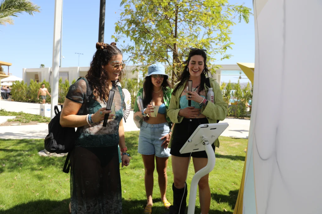 Three women laughing and enjoying drinks next to a photo booth at an outdoor event.