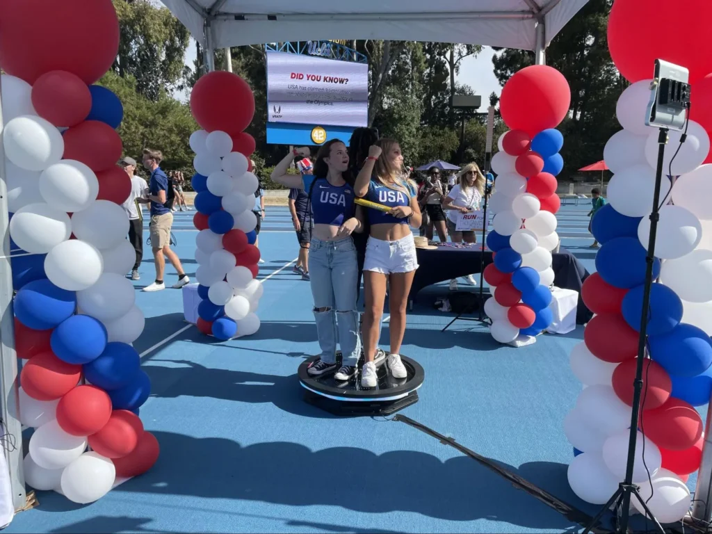 Two women standing on 360 photo booth platform at an outdoor patriotic-themed event.