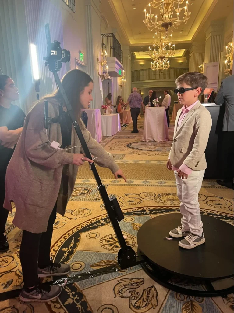 Young boy in sunglasses posing on 360 photo booth at elegant indoor event.