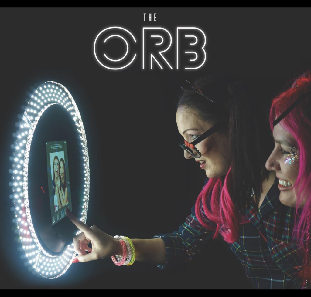 Two women interacting with a digital photo booth called The Orb, surrounded by glowing lights.