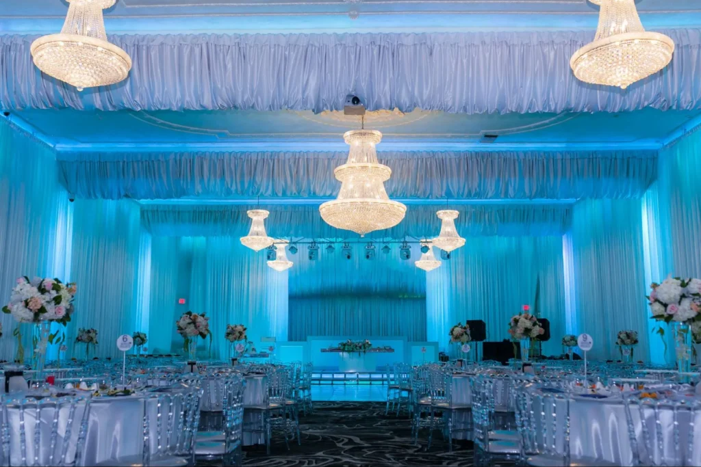 Blue-lit wedding hall with elegant chandeliers and table settings.