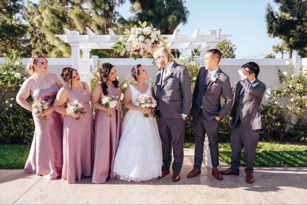 Bridal party under a floral archway with blush dresses and smiles.