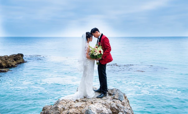 Bride and groom standing on seaside rock with bouquet.