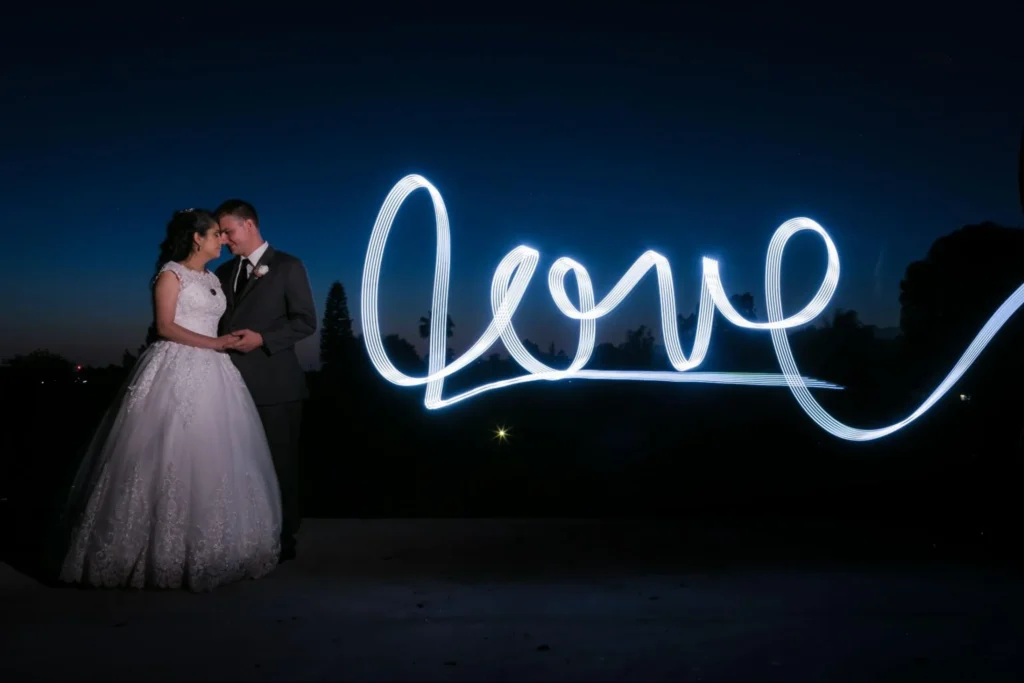 Bride and groom under 'Love' neon light at night.