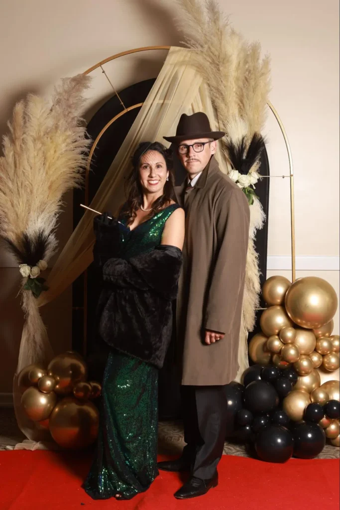 Elegant couple in vintage attire posing on a red carpet with gold and black decorations.