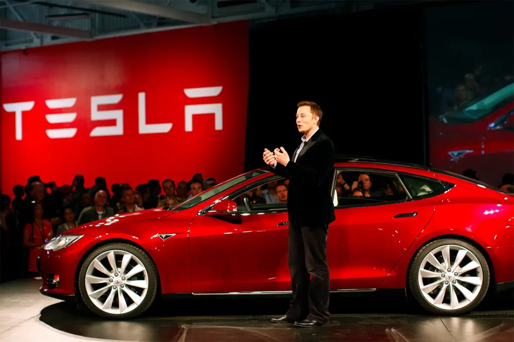 Elon Musk presenting in front of a red Tesla car during a corporate event with a Tesla logo backdrop.