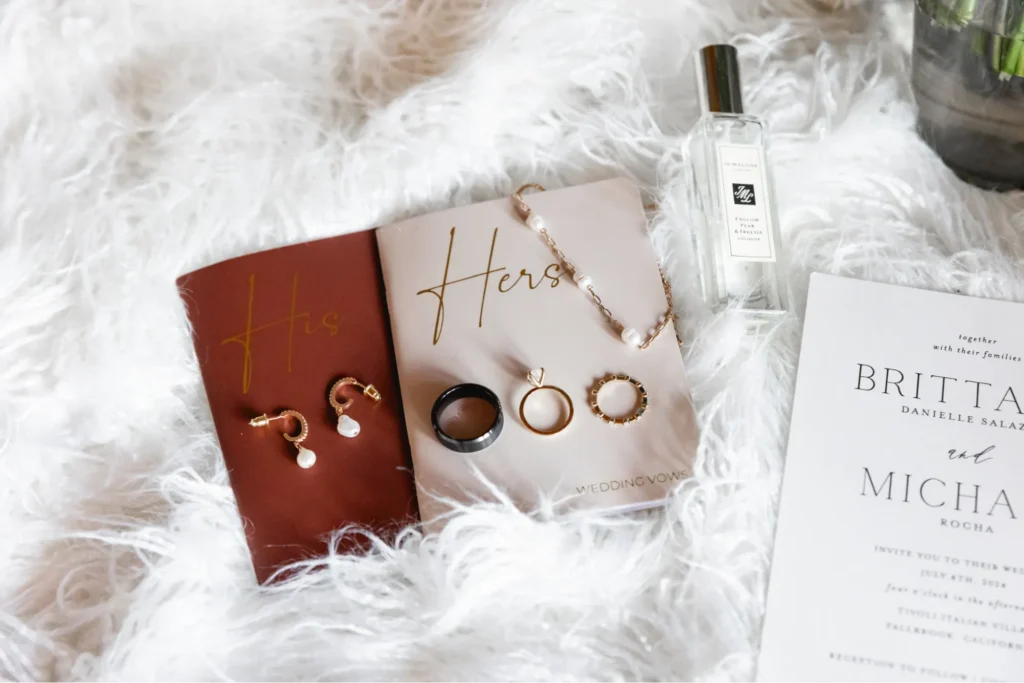 Flat lay of wedding rings, vow books, and perfume on a soft white background.