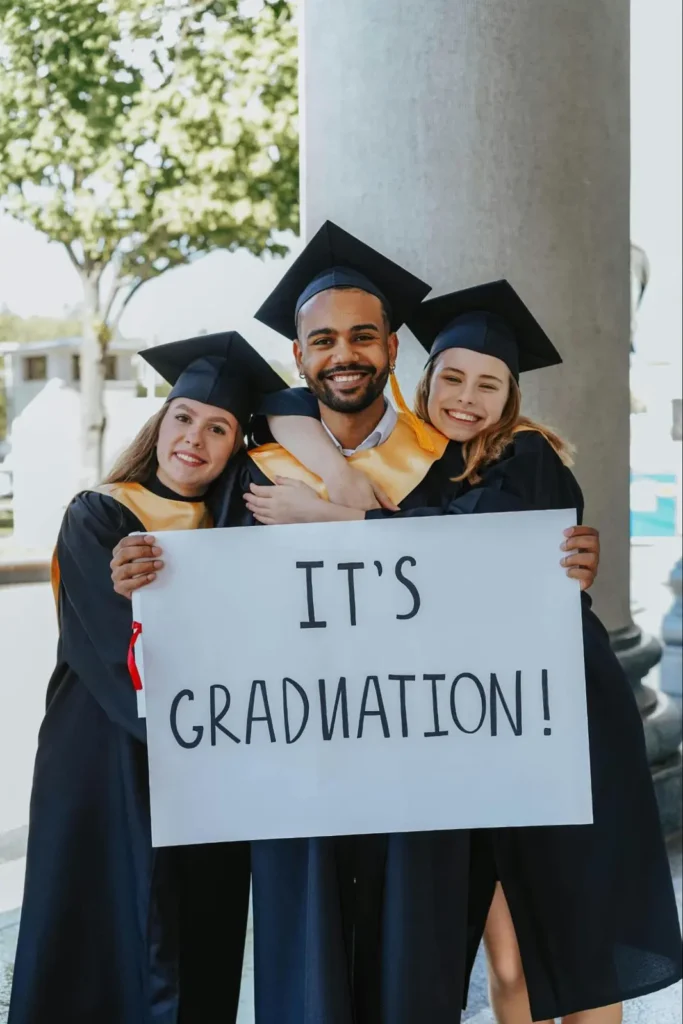 Group of happy graduates holding a sign that reads It's Graduation! in a celebratory outdoor setting.