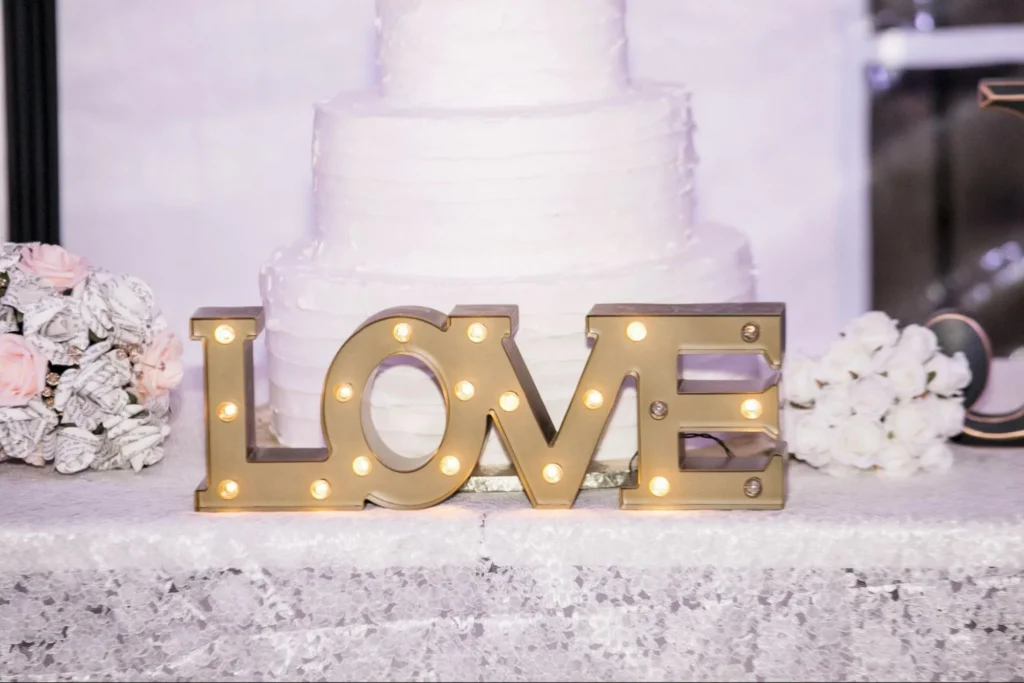 Illuminated 'LOVE' sign in front of a white wedding cake.