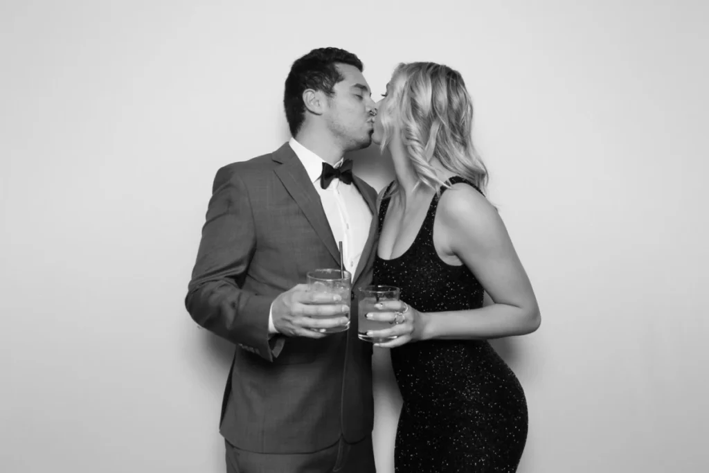 Romantic photo booth kiss pose at formal event, couple in suit and sparkly dress holding drinks, black and white