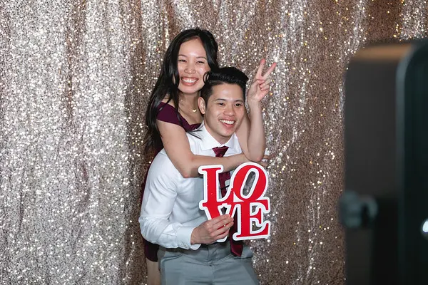 A smiling couple poses in front of a glittery backdrop. The man holds a LOVE sign, and the woman makes a peace sign.