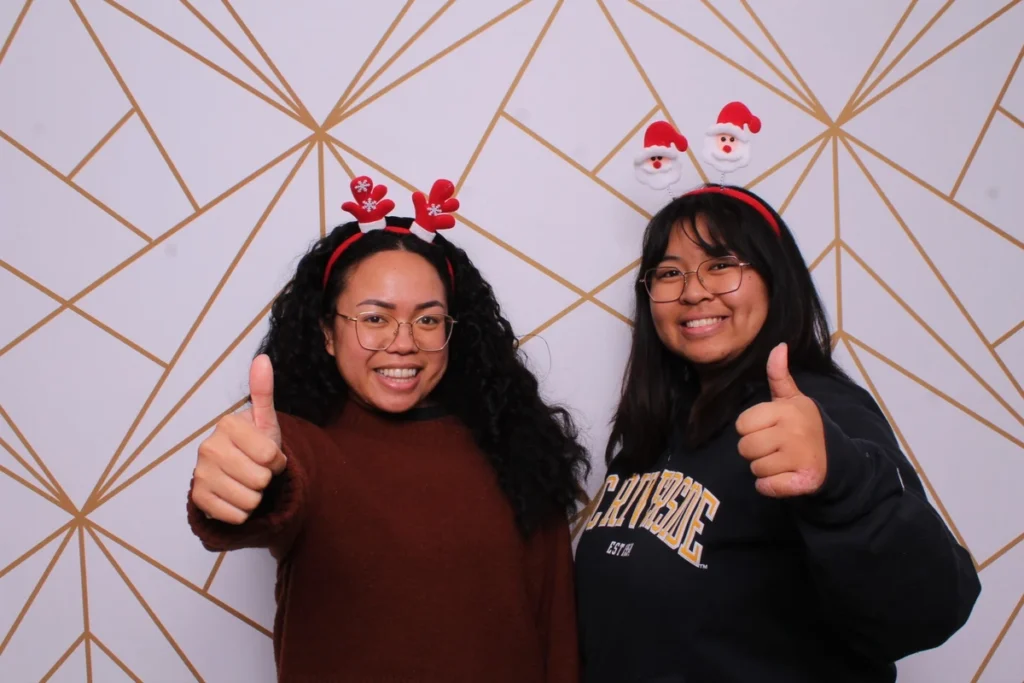 Christmas photo booth pose with Santa headbands, friends giving thumbs up against geometric backdrop, casual holiday style