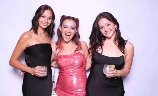 Friends striking fun photo booth pose at formal event, wearing black and pink dresses, holding cocktails with natural smiles