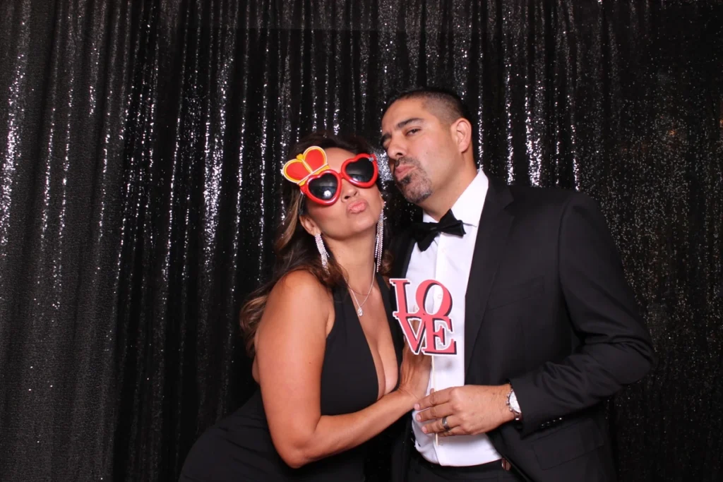 Glamorous photo booth pose with fun props - couple in formal wear against sparkly backdrop, heart sunglasses and LOVE sign