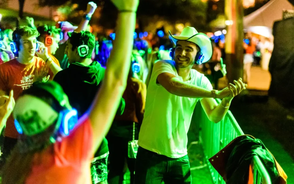 Man in cowboy hat line dancing at outdoor silent disco event, surrounded by participants with glowing headphones