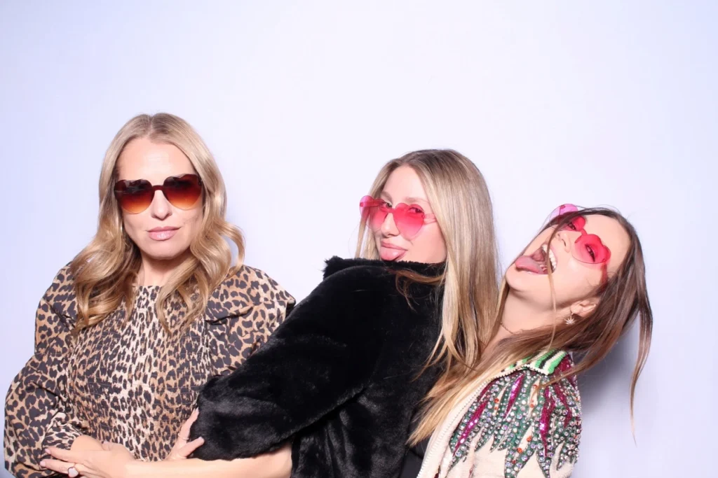 Three women wearing sunglasses striking fun poses in photo booth, displaying trendy group photo style