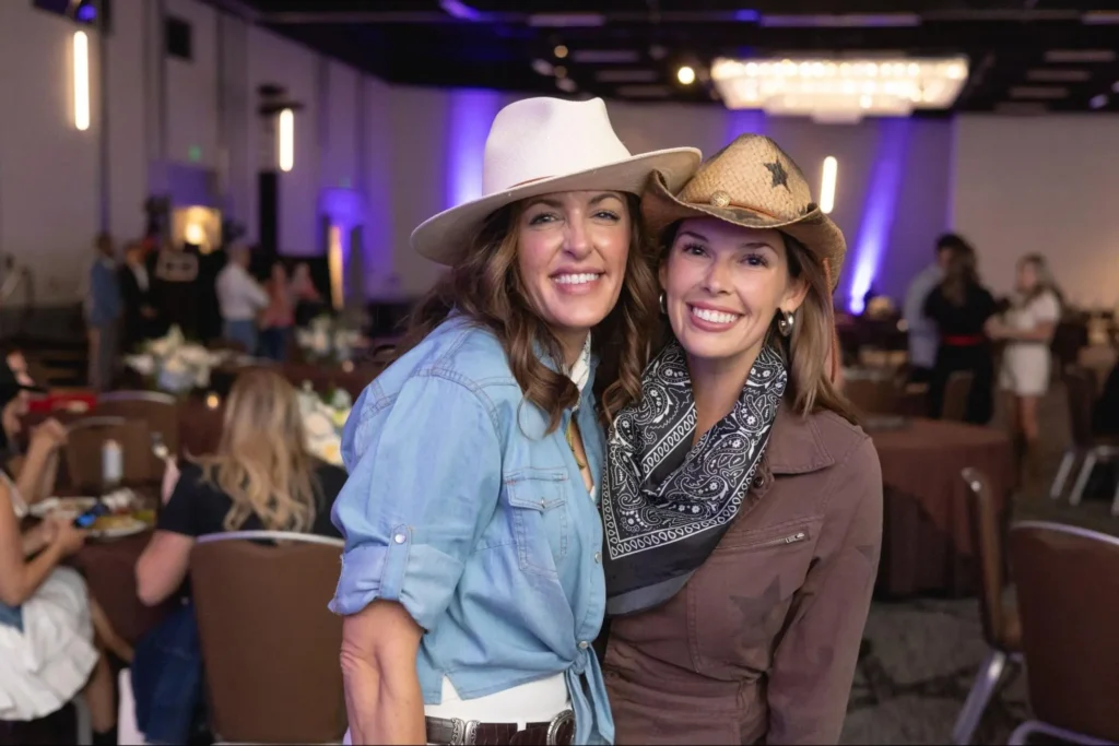 Two women in western attire with cowboy hats posing at a country-themed photo booth, wearing denim and bandanas