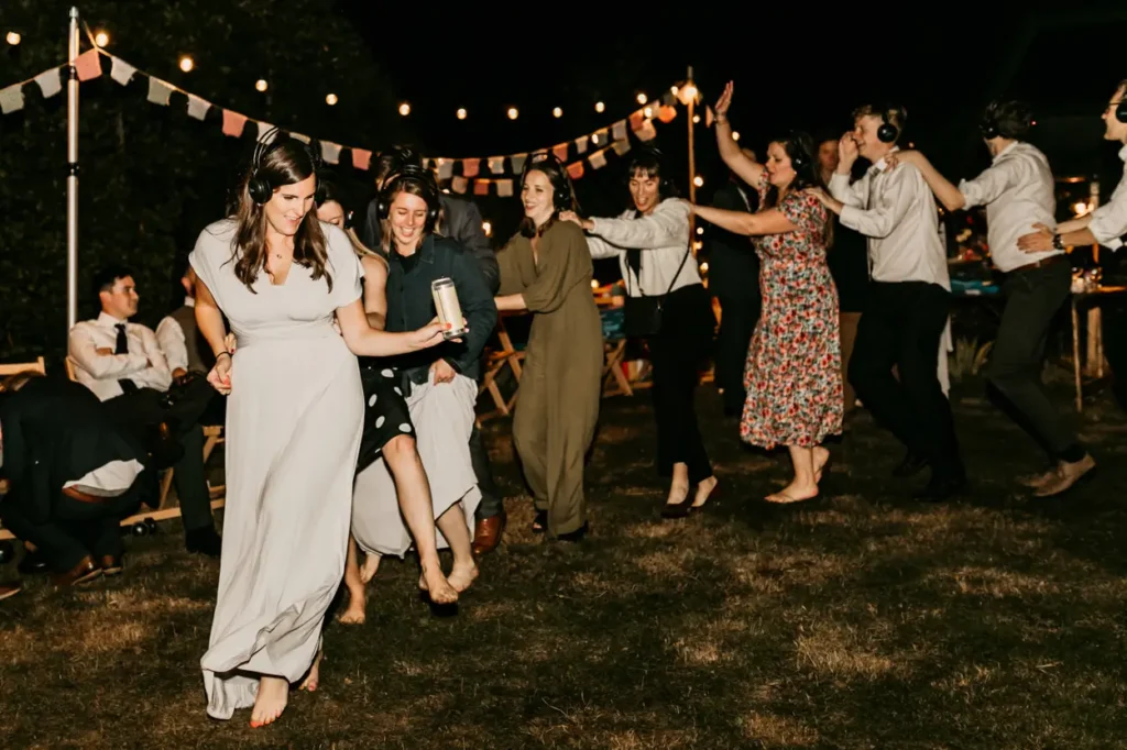 Wedding guests dancing with wireless headphones at outdoor silent disco, under string lights and bunting decorations