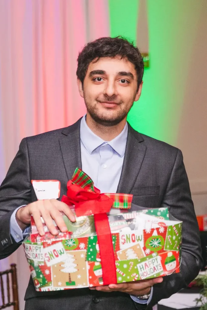 Young man in grey suit smiling while holding wrapped holiday gift with red bow at festive event