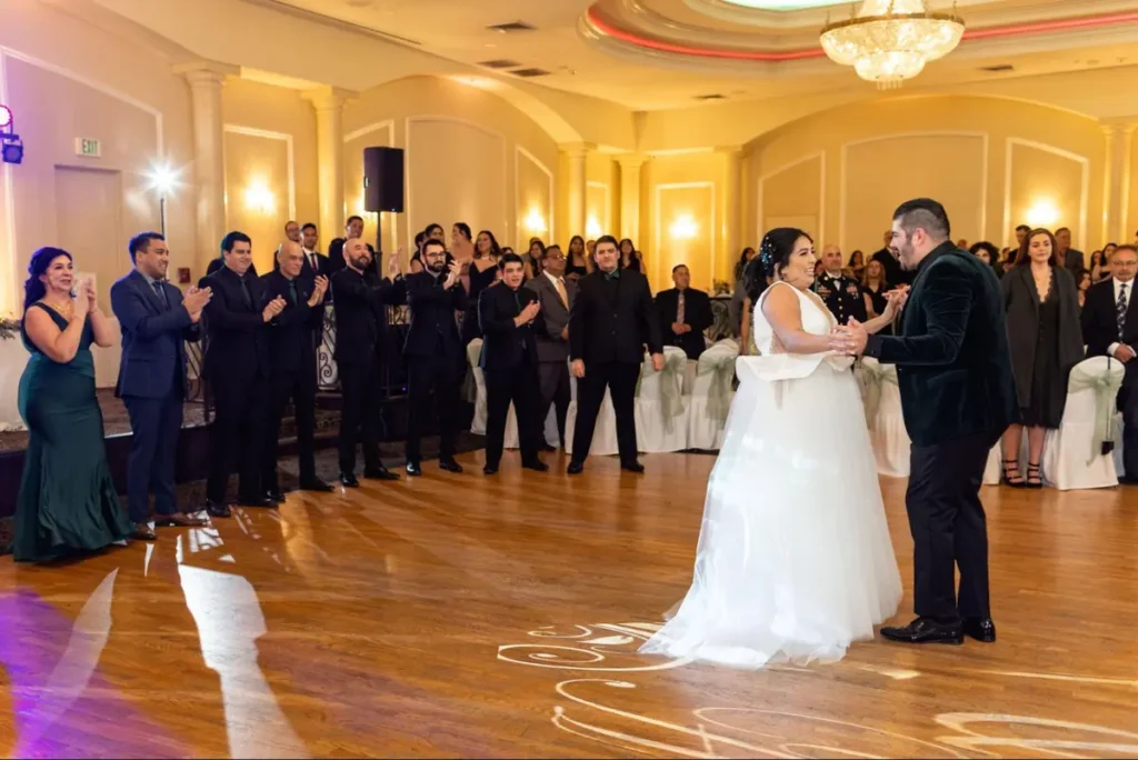 Bride and groom sharing first dance on elegant wooden dance floor with monogram and guests watching