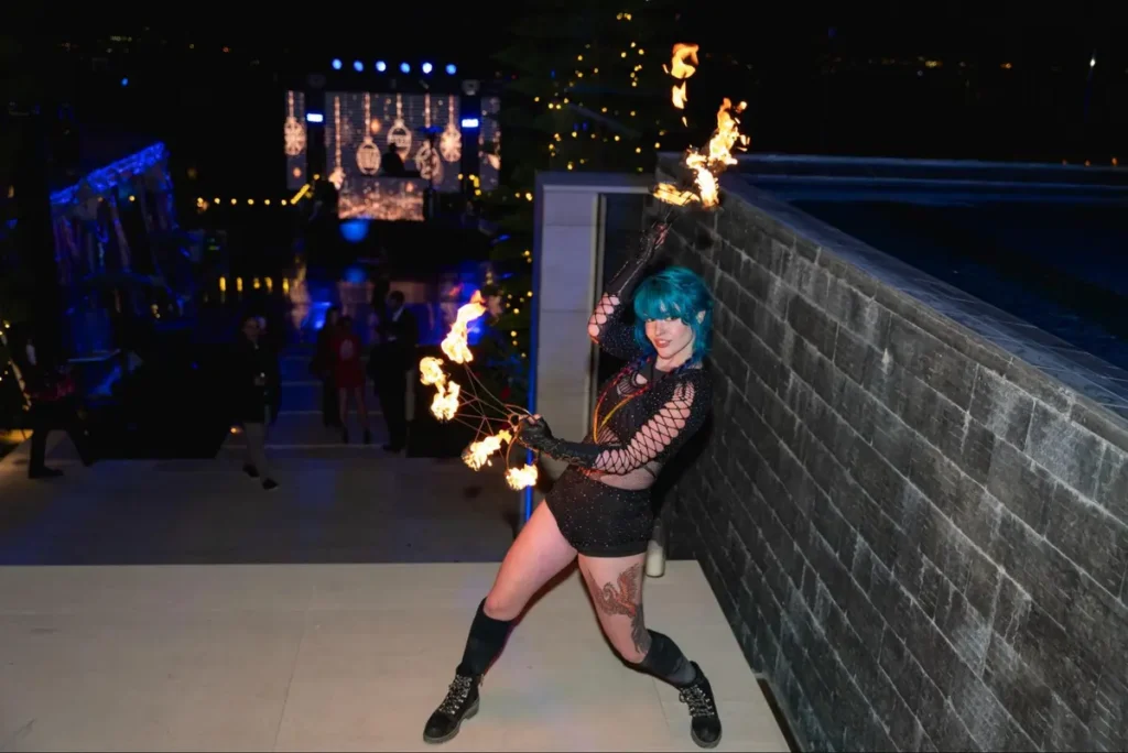 Fire performer with blue hair entertaining guests at a nighttime event with flames illuminating the outdoor venue