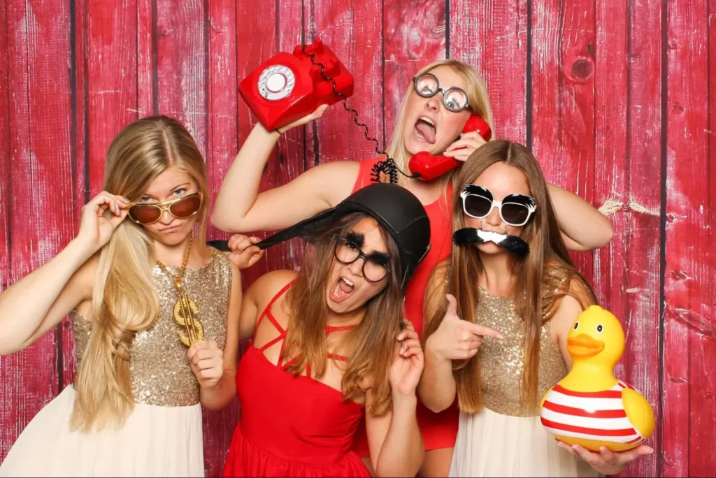 Group of friends posing with fun props in photo booth with red wooden backdrop, ideal for budget-friendly party rentals