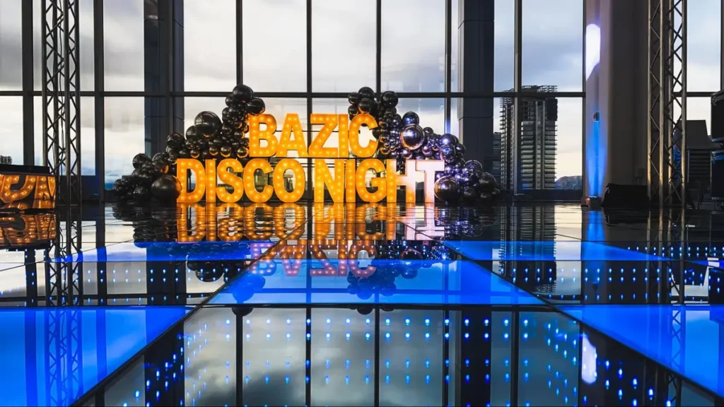 Illuminated 'BAZIC DISCO NIGHT' sign with black balloon arch and reflective blue floor creating dramatic event atmosphere