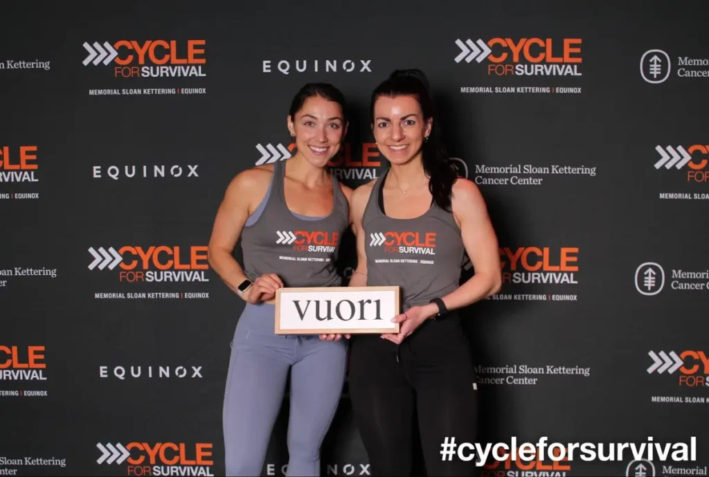 Two participants in gray tanks at Cycle for Survival charity event by Memorial Sloan Kettering and Equinox, holding Vuori sign