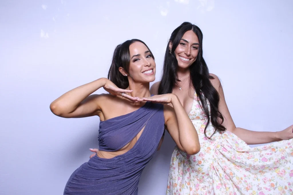 Two women posing together in a photo booth, one in purple dress making hand gesture, other in floral dress against white background