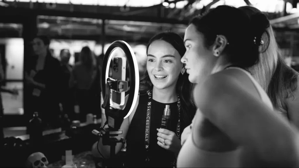 Two women using a ring light selfie booth with smartphone mount at an event, black and white photography