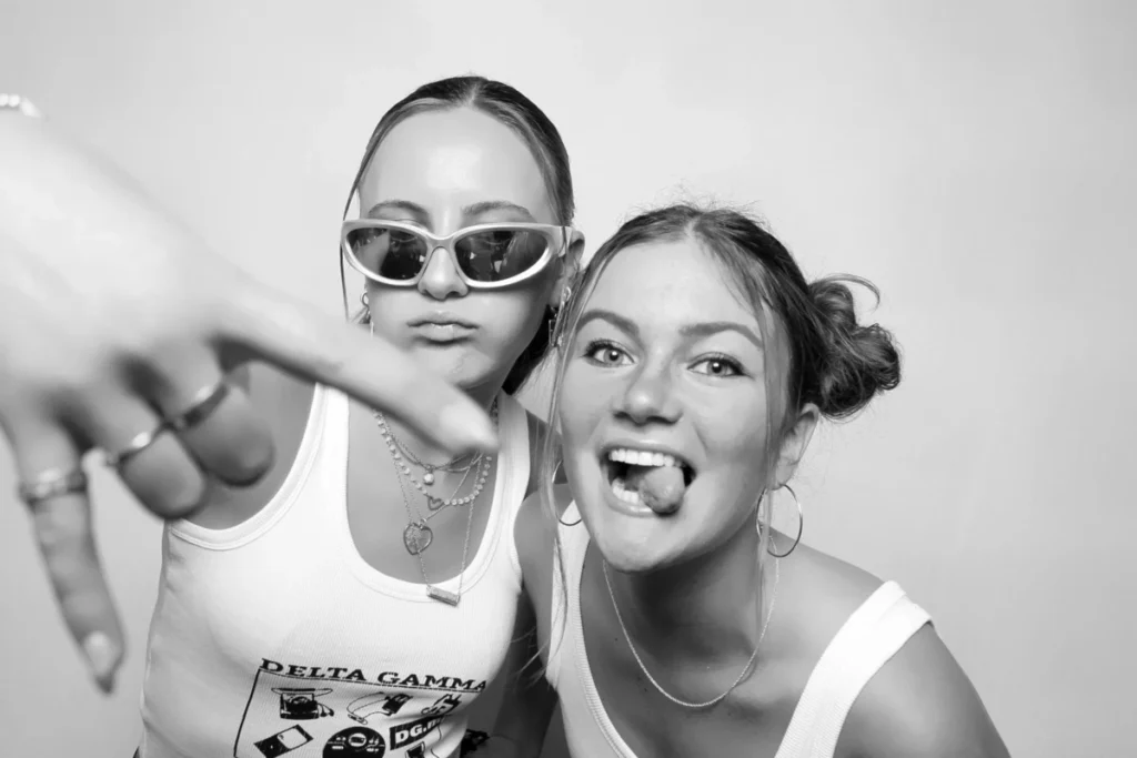 Two young women posing for a black and white photo booth picture, one wearing sunglasses making a peace sign
