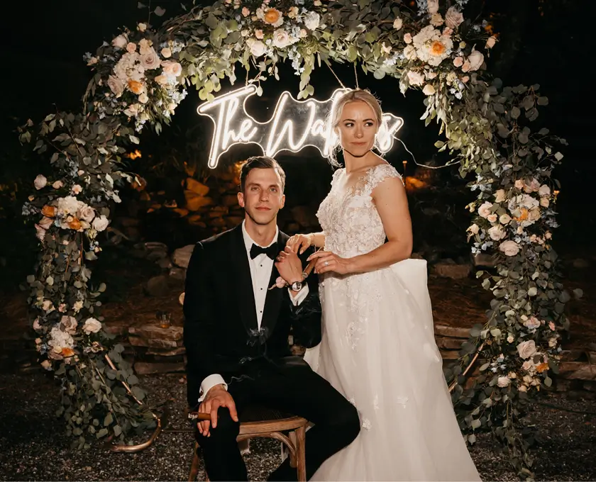 Wedding photo booth with floral arch and neon 'The Waits' sign, creating elegant backdrop for newlywed couple