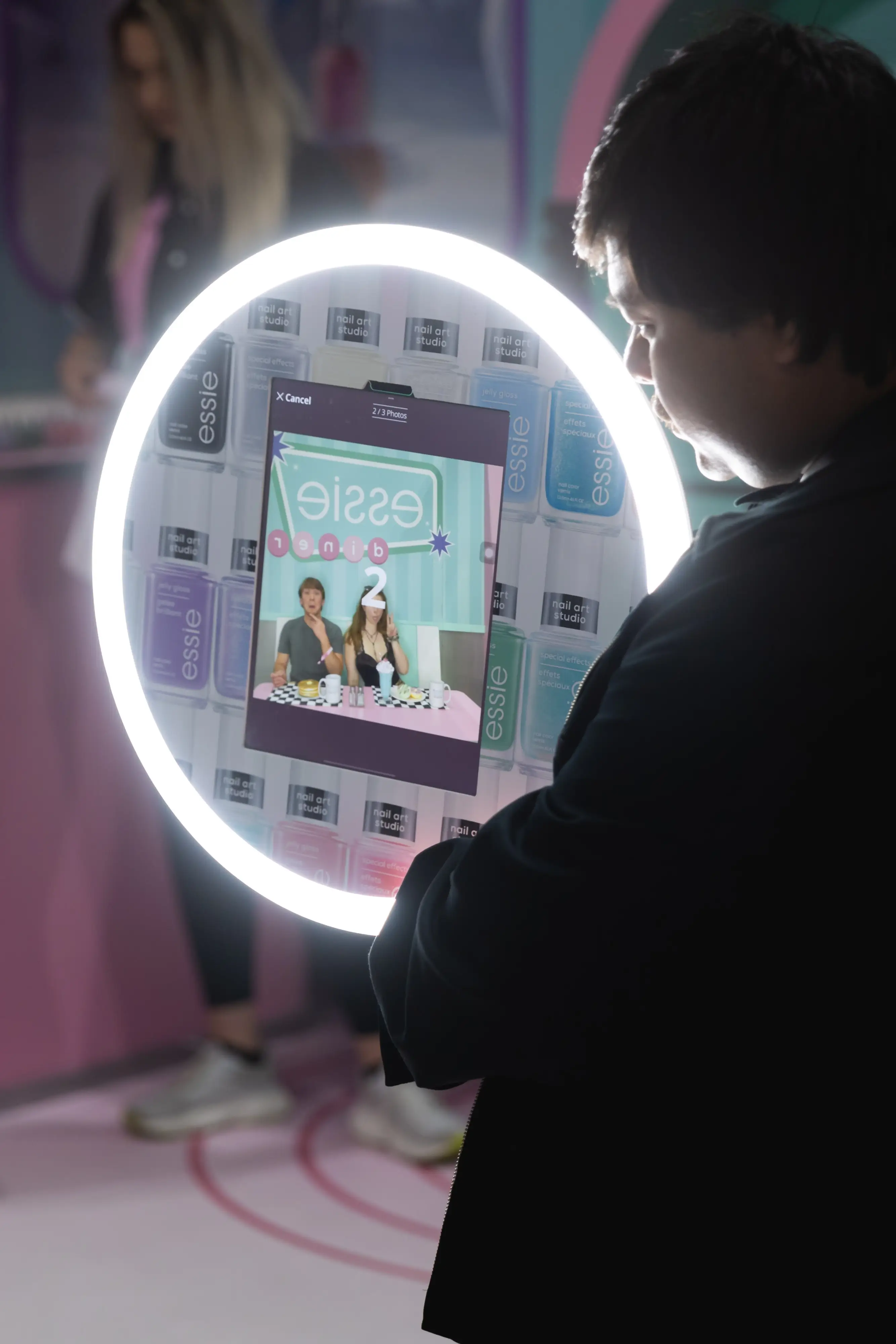 Attendee interacting with Essie's Roamer Booth at DragCon, viewing a retro diner-themed photo on the illuminated display against a backdrop of Essie nail polish product