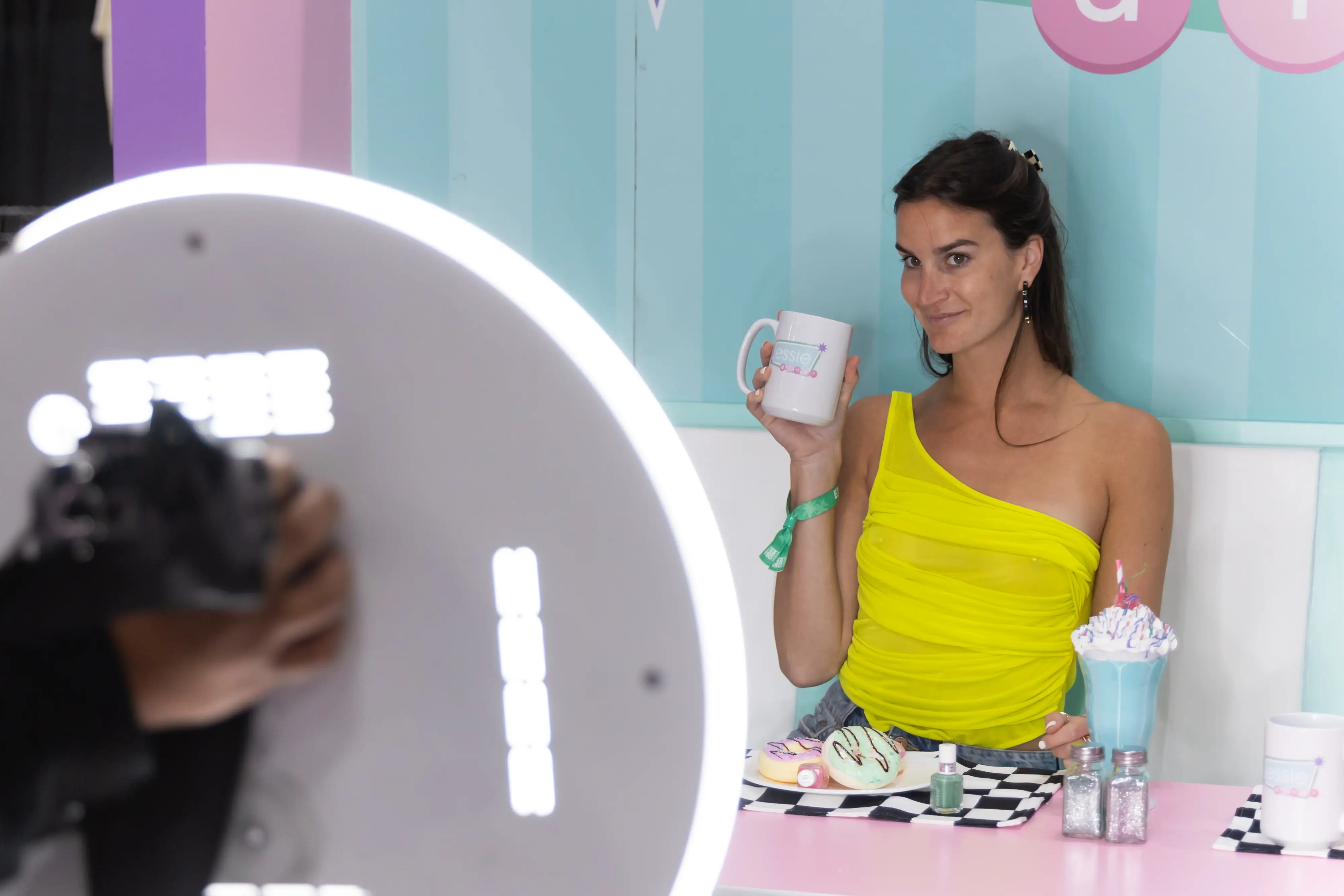Woman in bright yellow top enjoying the Essie diner experience at DragCon, posing with themed mug and treats while the Roamer Booth's ring light frames the scene